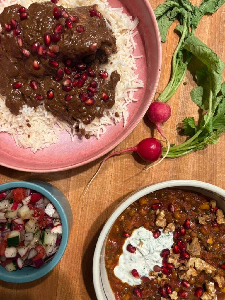 A pink high-sided plate containing basmati rice topped with a dark walnut sauced chicken and pomegranate seeds, a small blue bowl of salad shirazi, a light blue bowl of walnut and pomegranate soup topped with yogurt and additional pomegranate seeds and walnuts, and fresh radishes.