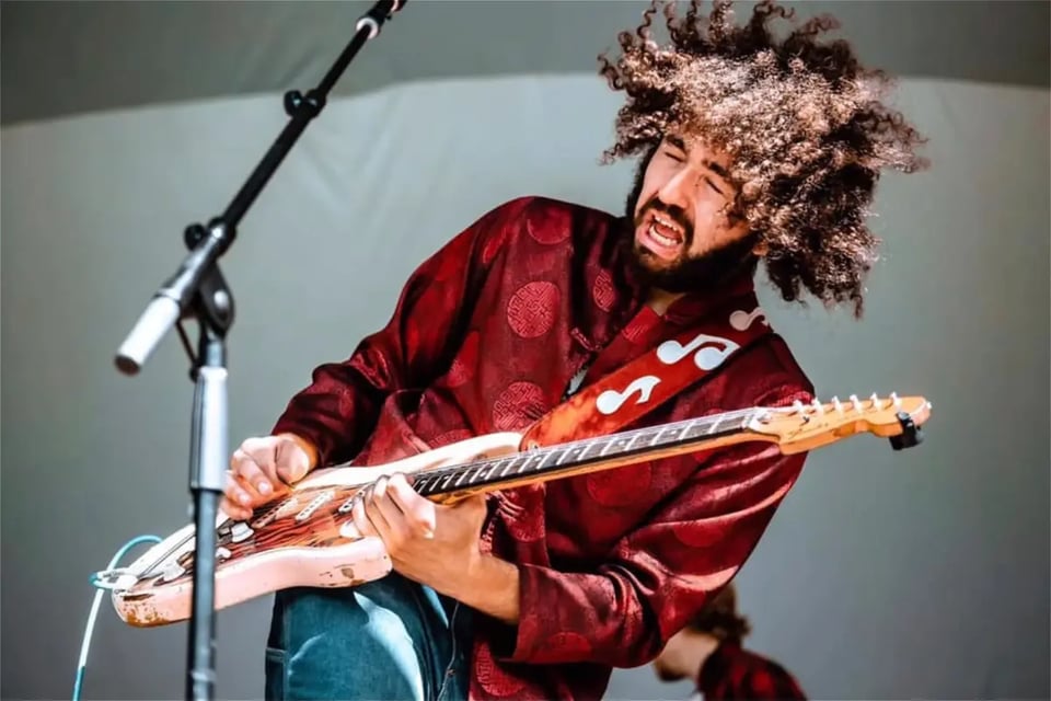 A man with a voluminous afro wearing a red smoking jacket and jeans plays a pink Fender Stratocaster guitar with his eyes closed and mouth open.