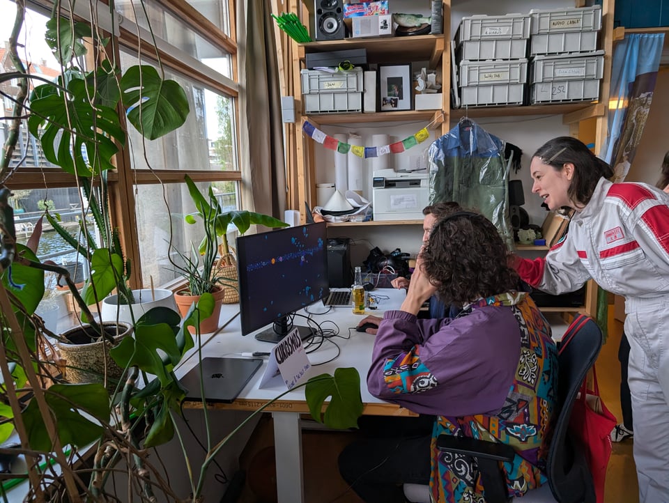 Three people play a video game, their desk framed by a large Monstera plant, and shelves full of colourful boxes and flags.