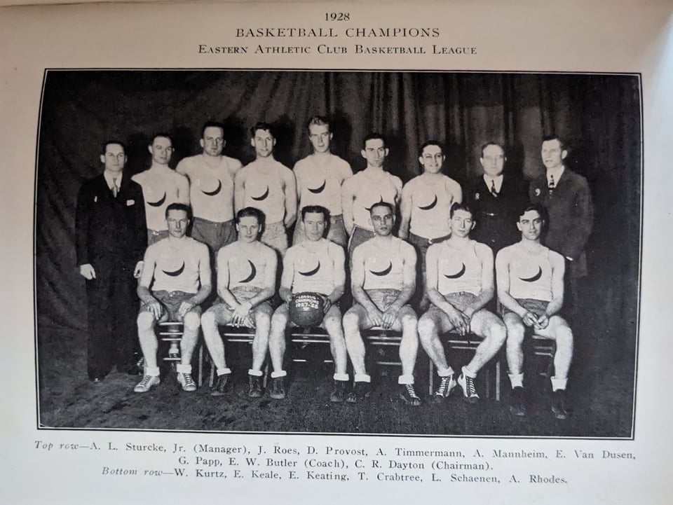 The club basketball team, wearing white shirts with dark crescent moons at the center, posing with their coaches (one row sitting, one standing) as one of their players holds a game ball from their championship.