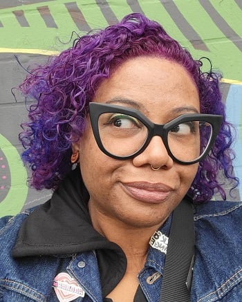 Outdoor selfie. I'm wearing large black cat eye glasses and looking to the side. My short curly hair is fuchsia, violet, and blue.