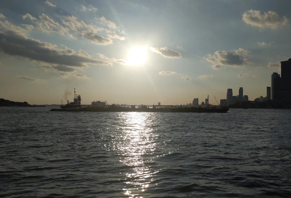 A tugboat pushing a barge up the East River between Brooklyn and Manhattan (edge Manhattan Skyline visible in the background) beteanth the setting and very bright Sun.