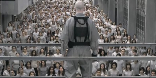 A still from the first season of 3 Percent on Netflix, showing a bald man wearing a harness, standing on a walkway looking down at a sea of young people wearing identical white clothes