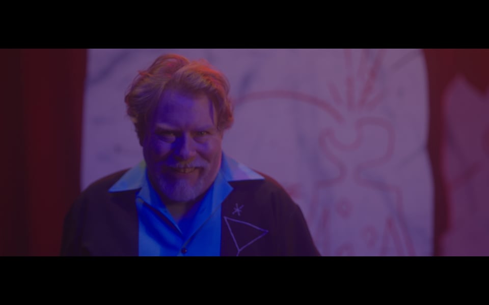 A frame from the film Dead Media. The actor, Sam Landman, grins at the camera, strange cave art behind him