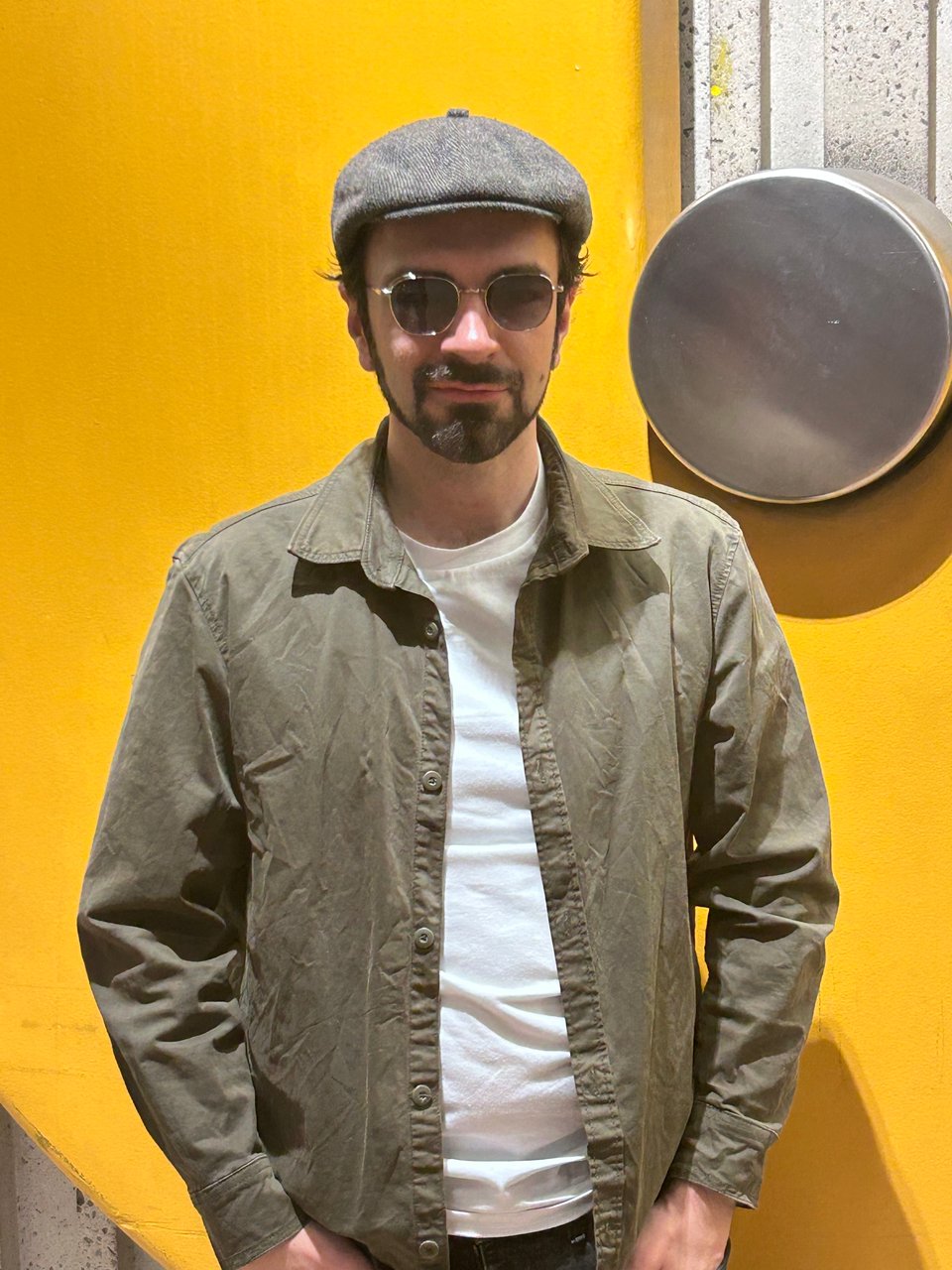 Photo of Matthew in a flat cap and olive jacket wearing a pair of aviator-like shades beyond an orange and stainless steel disk in the Montreal metro system.