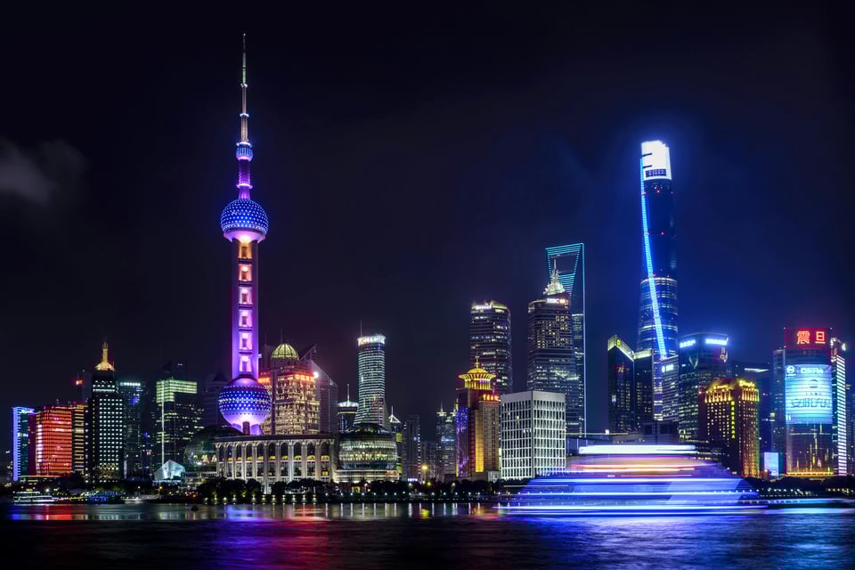 A picture of Shanghai at night. There are several large towers looming into the dark sky, and they are luminously lit up, casting a glow all around them and onto the water in front of the city