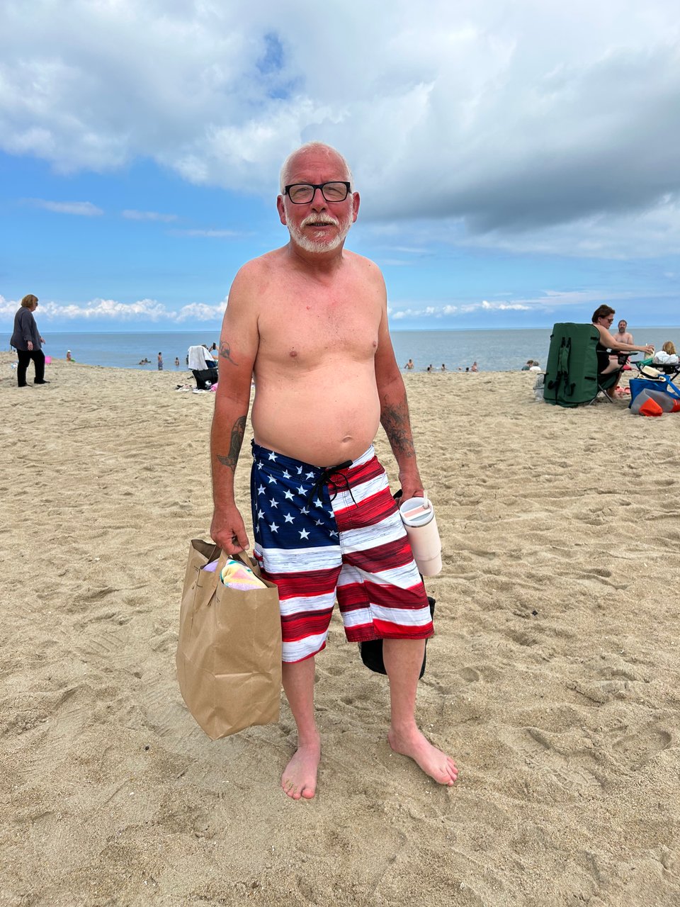 60ish White man shirtless with white beard and mustache, wearing U.S. flag swimming trunks and holding a shopping bag of towels and supplies, standing on sandy beach with the ocean behind him.