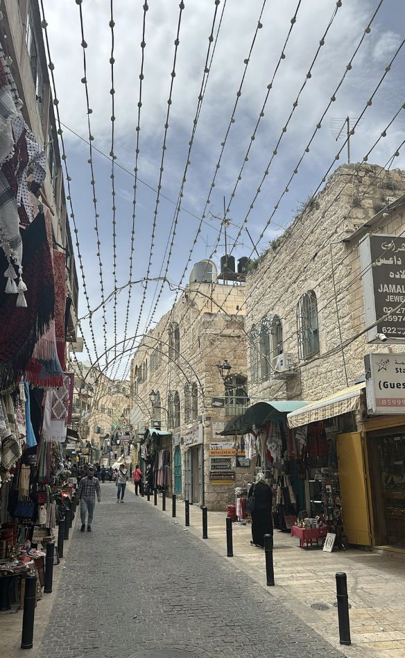 A long cobblestone street with just a few people, shops on the sides of the street. Lights strung atop the walkway.et