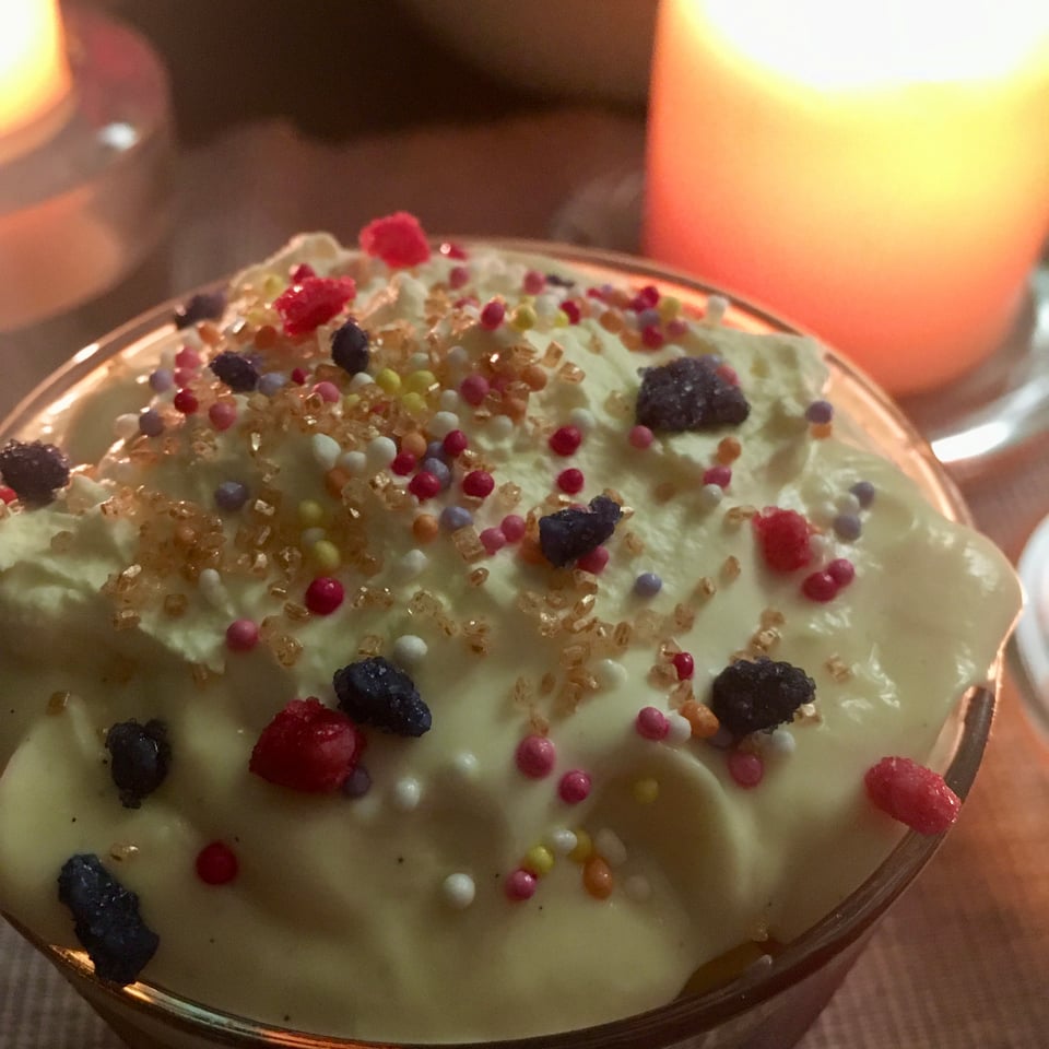 A glass filled with trifle. The top is softly whipped cream scattered with many different kinds of sugar sprinkles. There's a fat beeswax candle glowing in the background.