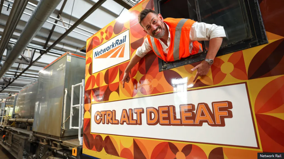 a photo of a train driver leaning out teh window of a leaf cleaning train. the driver is wearing a high-vis vest. The train is brightly cloured with various shades of orange shapes. The train name is printed on the side in orange and brown text - "CTRL ALT DELEAF"