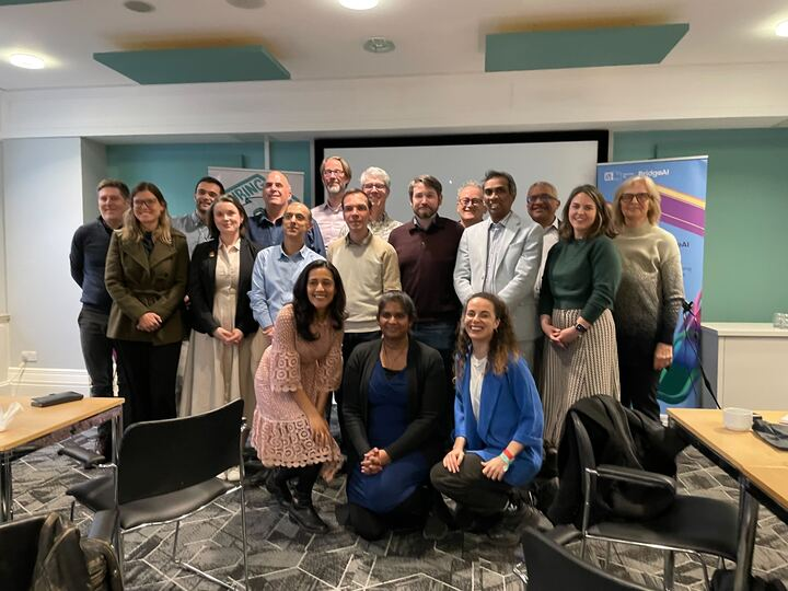 Image of 18 people standing in front of a room in professional clothing. There are chairs, tables, and pieces of paper on the desk.