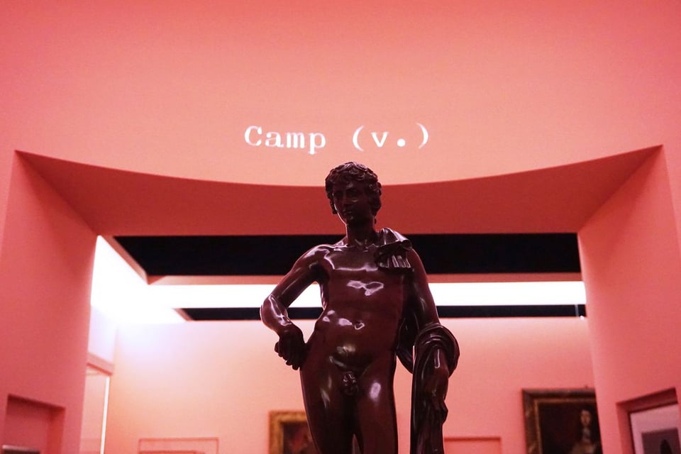 A nude male statue with its hand on its hip, in a room with pink walls and the word "Camp (v.)" written on top of the archway in white letters