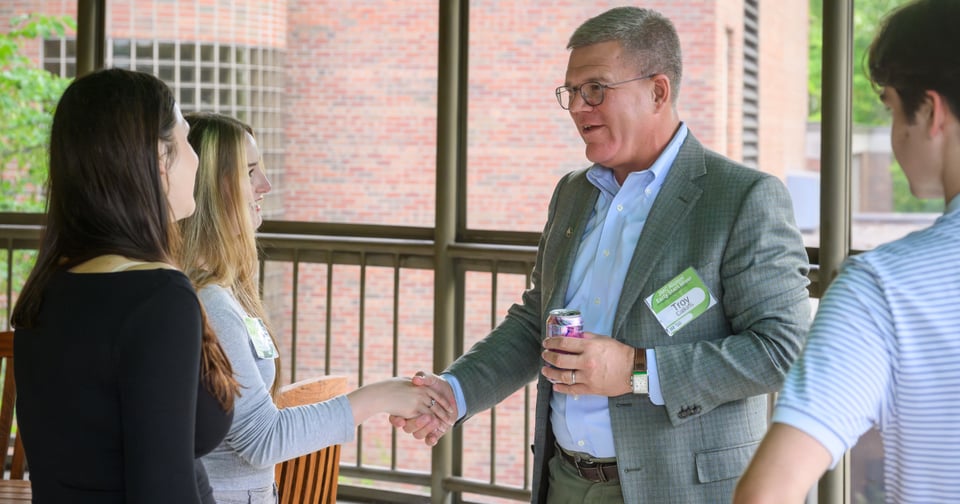 A JMC alumnus wearing a grey blazer and blue collared shirt, with arm extended shaking hands with a JMC student.