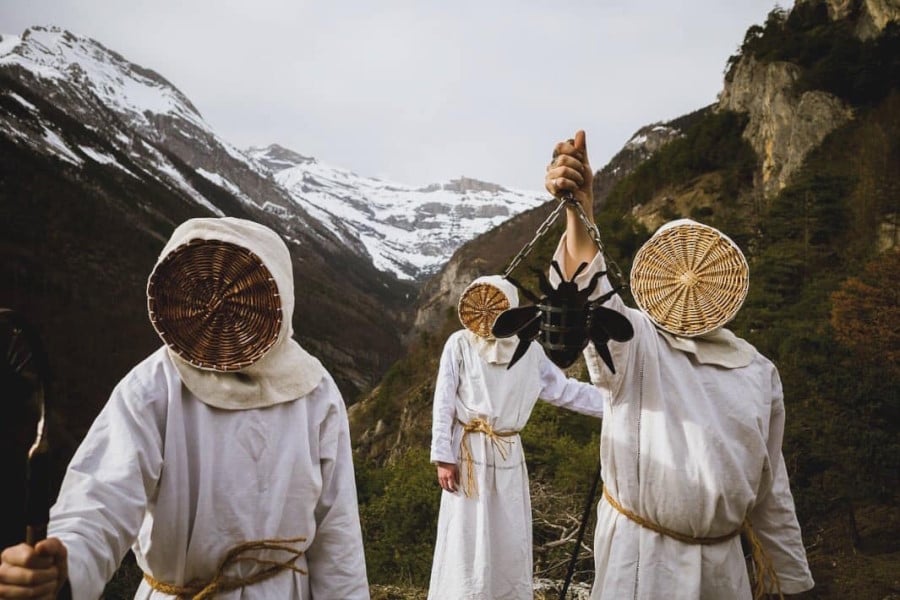 Three men dressed in white cloaks with ropes on the waist. They wear the top of wicker baskets as masks. The man on the left is holding a bee hanger. The outfit is based on medieval beekeeper apparel. They are posing on top of Swiss mountains, some of them with snow in their peaks.