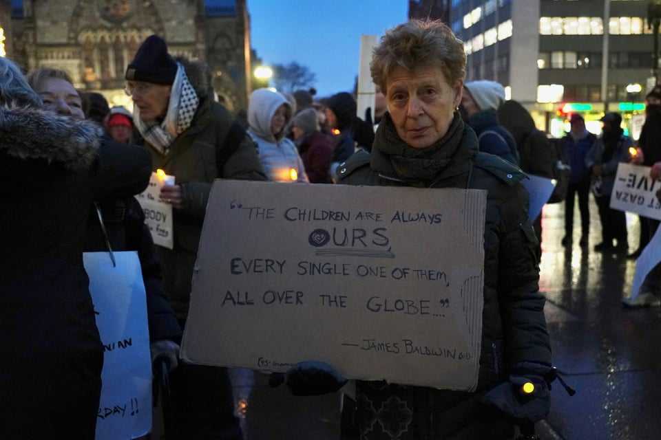 A protestor holds a cardboard sign in a larger group of people. The sign is a James Baldwin quote, "The children are always ours, every single one of them, all over the globe." The word "OURS" is larger and underlined twice.