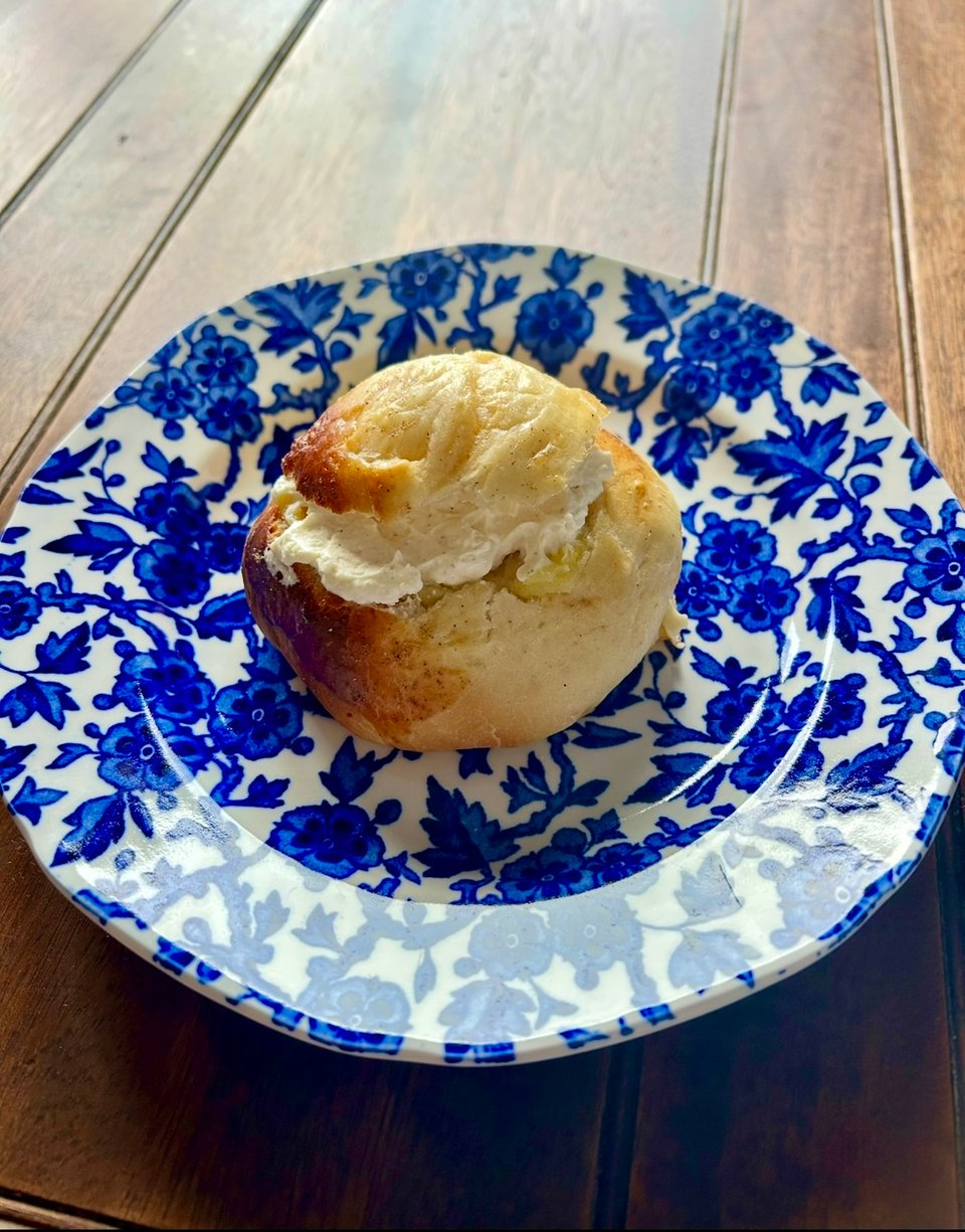 A round Swedish-style semla bun, with cream peeking near the top is on a blue and white plate, sitting on a dark brown antique mahogany table. Image by Rowan Ambrose