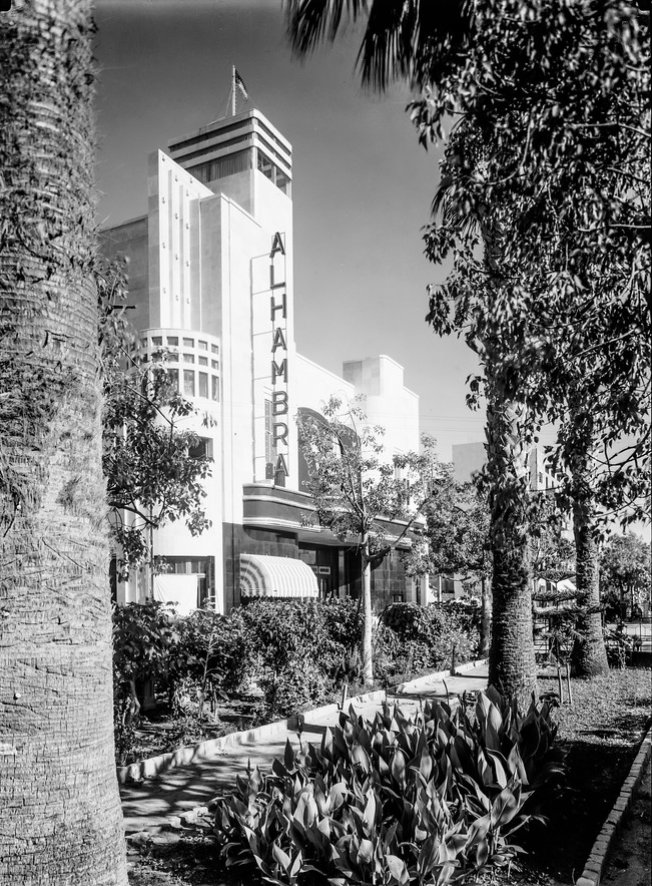 A tree-lined street with a movie theatre with the name ALHAMBRA