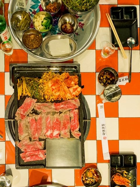 A tray of banchan sits above slices of beef cooking on a griddle top on a red and white tiled table