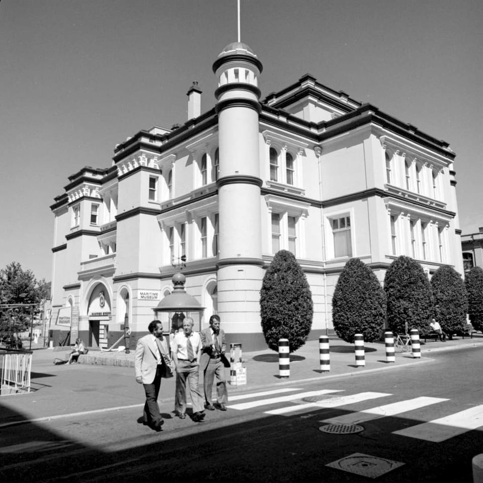 Black and white photo. 3 men in suits cross the street in front of the Maritime Museum or Old Court House building