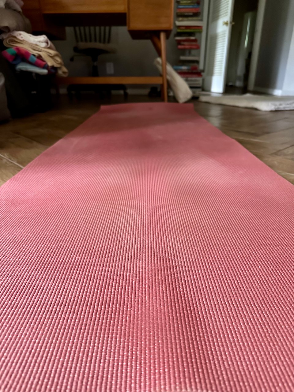 Photo of yoga mat, taken near floor level, so it’s receding into the back of the frame, leading to a desk, a bookshelf, and a canvas tote bag