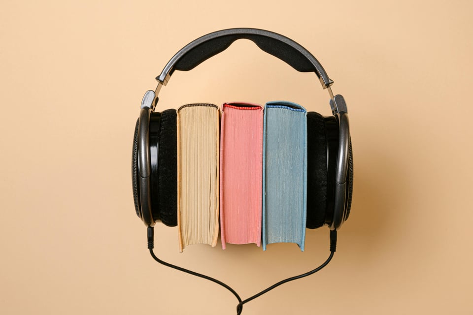 Image of a pair of headphones wrapped around three books that are yellow, red/pink, and blue.