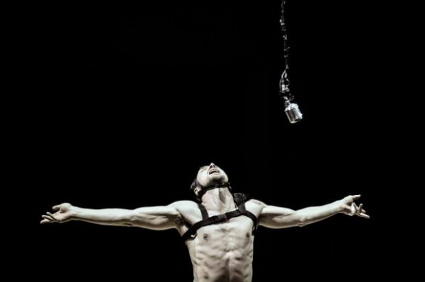 Lars Eidinger in Thomas Ostermeier's Richard III. He is shirtless except for a bondage vest, and has his arms out and his head back. An old-timey microphone dangles above him.