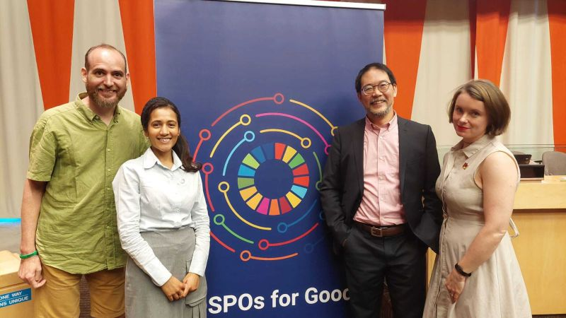David Perez Suarez, Malvika, Neil Chue Hong and Arielle standing next to a big dark navy background with the UN's sustainable development goals symbol, a multicolored ring with different multicolored lines encircling it