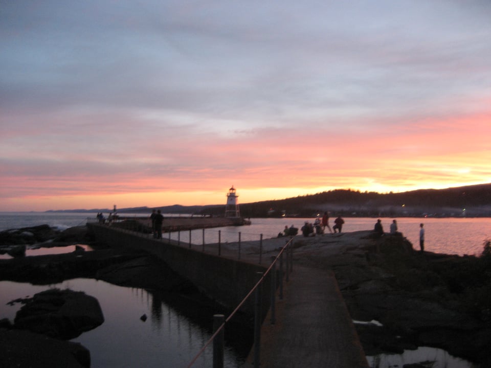 A harbor breakwater with a few people on it and a lighthouse on the end. It is sunset and the sky is red and gold by the horizon, and the water reflects it almost pink.