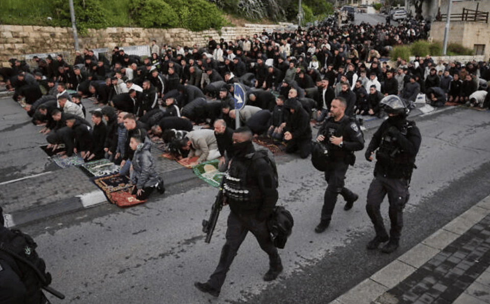 Photograph showing a very large group of people praying outdoors on a street, with several individuals kneeling on prayer rugs. Four police officers in black tactical gear walk past the worshippers
