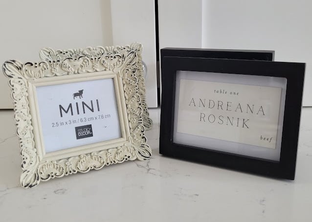 Four small frames on a white countertop. The two frames on the left, one in front of the other, are cream-colored and ornate. The two on the right, one in front of the other, are classic black solid plain frames.