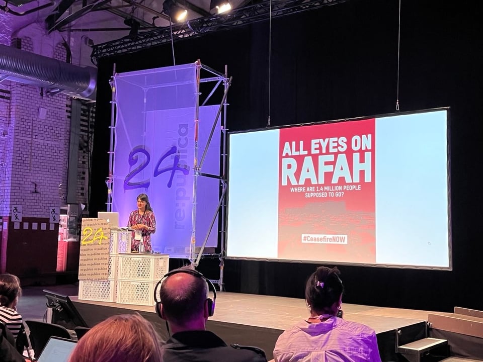 A stage at a conference, with a large screen showing a red image with the white text All Eyes on Rafah written on it, subtitle Where are 1.4 million people supposed to go. A hashtag Ceasefire now is visible at the bottom of the picture. An asian woman is standing to the right of the stage by a laptop, mid speech.