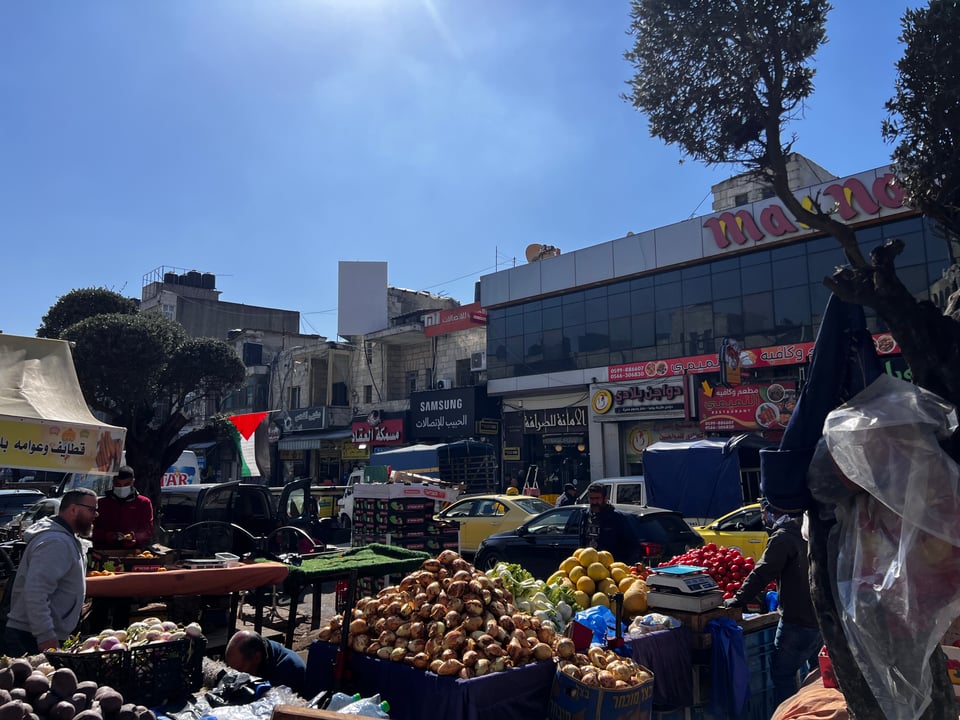 Produce for sail on a street, a Palestinian flag flying, and a large building on the far side of the street
