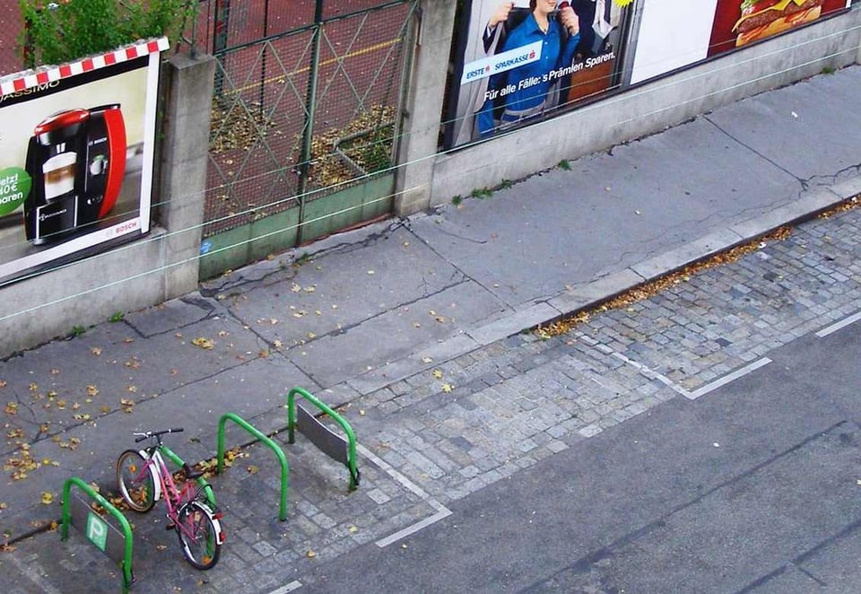 Foto de la calle hecha desde un edificio. Se ve un aparcamiento de bicis con una bici rosa.