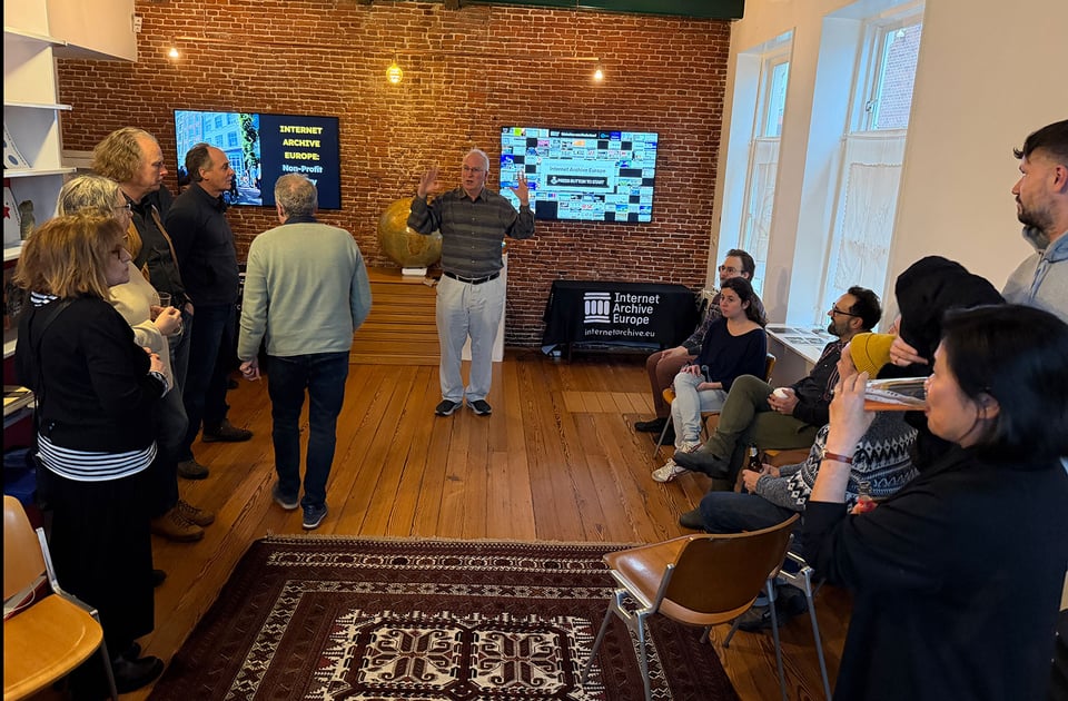 A man stands in front of a room with two screens at the far end. The audience of people stand around the edges of the room.
