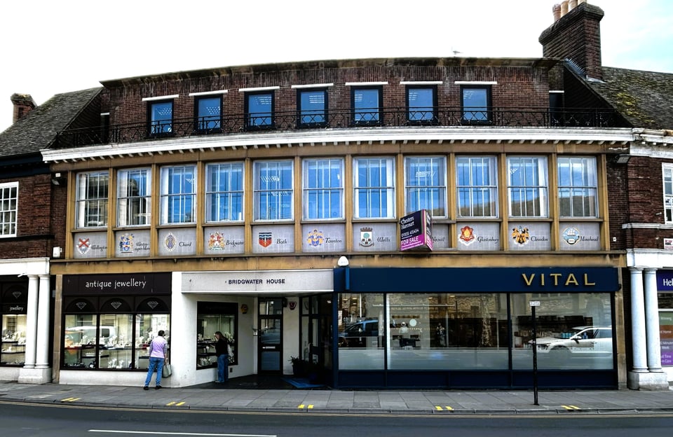 A very midcentury building, with retail on the ground floor and offices above. the upper storey has eleven large windows set into simple stone panels. Below the windows the rest of the panel is tiled with motifs for several towns in Somerset. A mansard roof with more windows looks like a later addition.