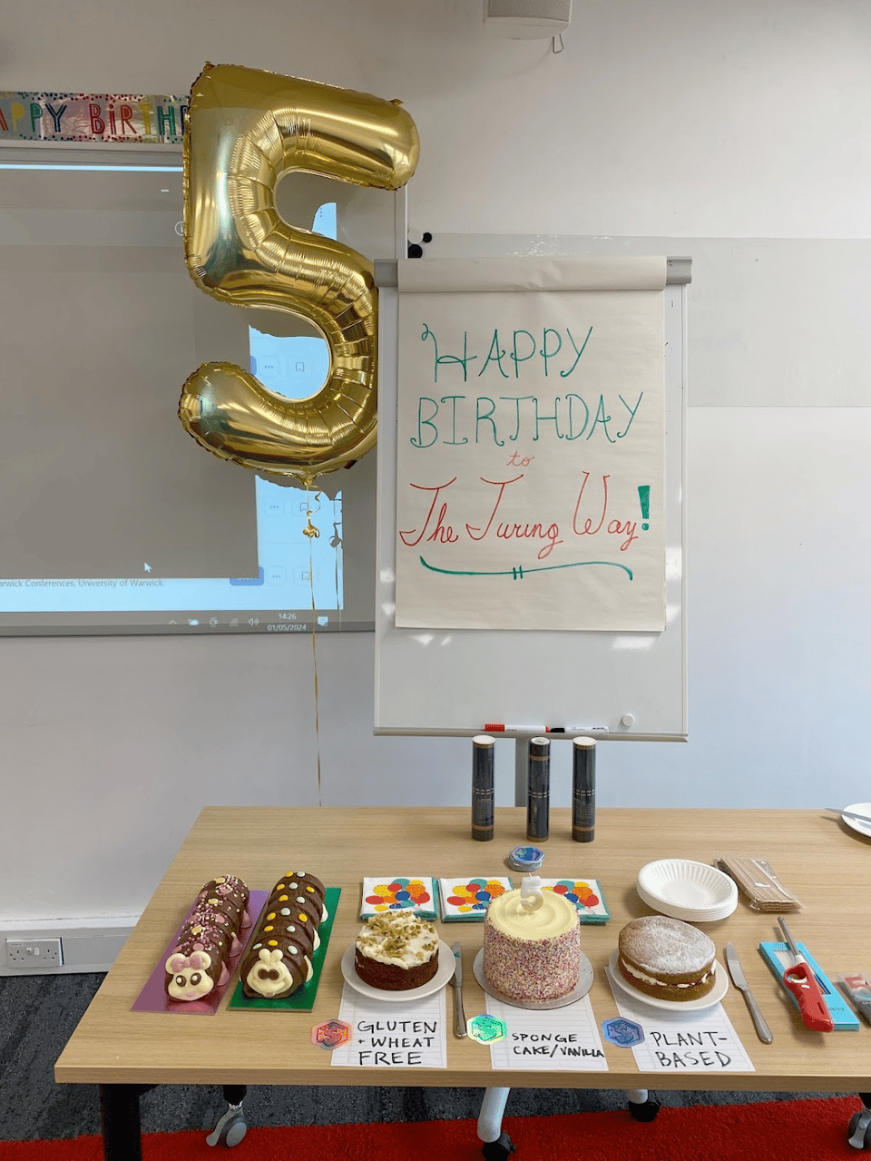 Alt: A festive scene celebrating _The Turing Way_’s birthday, featuring a golden balloon shaped like the number 5. Above it, birthday decorations peek out, and next to it stands a flipchart with "Happy Birthday to _The Turing Way_" written on it. In the foreground, a table with colorful cakes, including two in the shape of caterpillars — a beloved British treat!