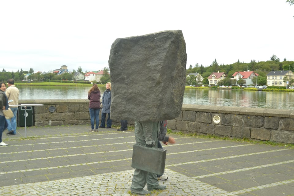 A statue of a man in a suit carrying a briefcase. His head and upper torso have been replaced with a large, square cut rock.