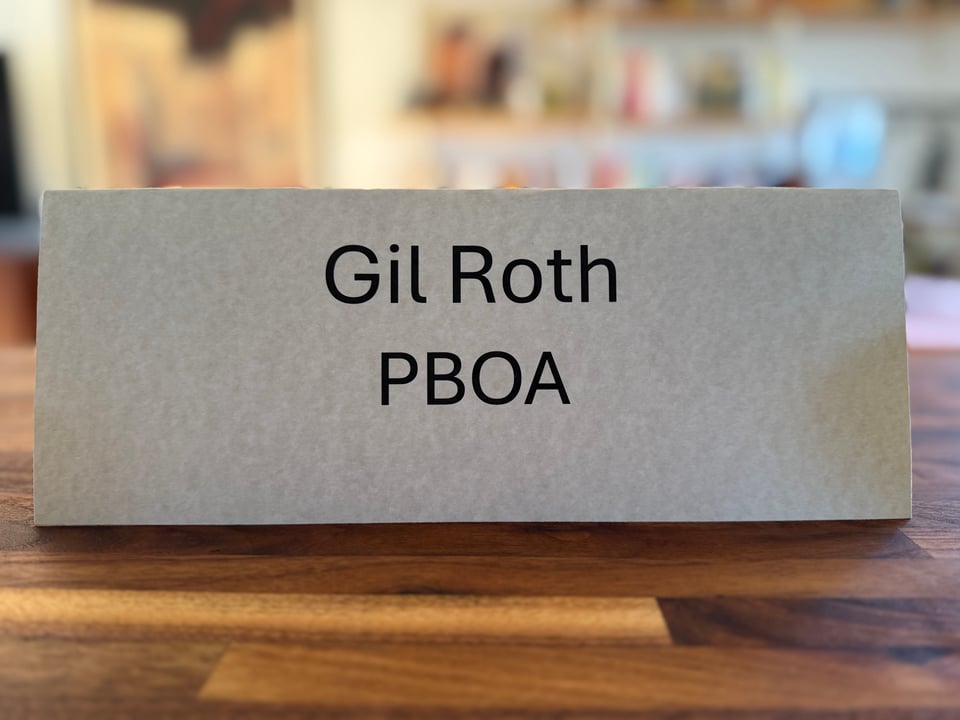 Off-white placard on a butcher-block table that reads, "Gil Roth • PBOA"