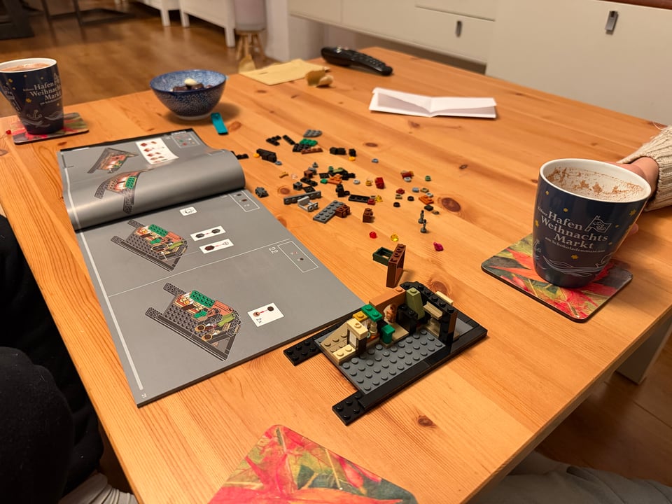 A coffee table is covered in legos with an instruction book open. The build has started, there are two cups of hot chocolate.