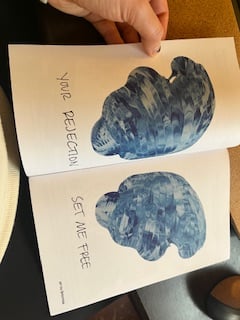Nola holding open a page of the Transness is Our Salve zine, with blue watery wing images and the words "your rejection set me free"