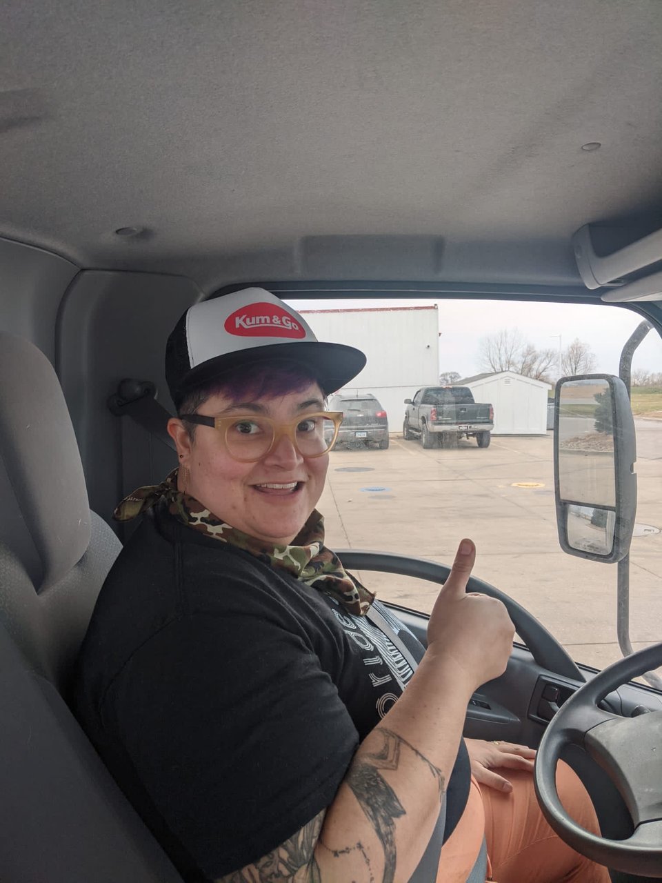 KCF in a truck cab smiling awkwardly and signaling thumbs up in a kum and go trucker hat