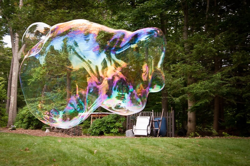 An enormous, irregularly shaped soap bubble floating above a green lawn with trees, a hed, and two wheelbarrows in the background