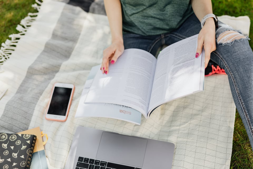 Image of a person, face not seen, opening a notebook while sitting on a blanket outside.
