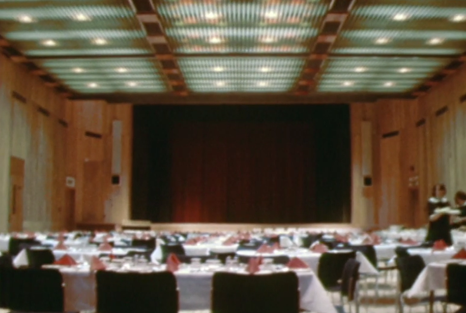 The same room set for a banquet with tables set with white clothes and pink napkins. The ceiling is lower, and now has lights set into aluminum cladding.