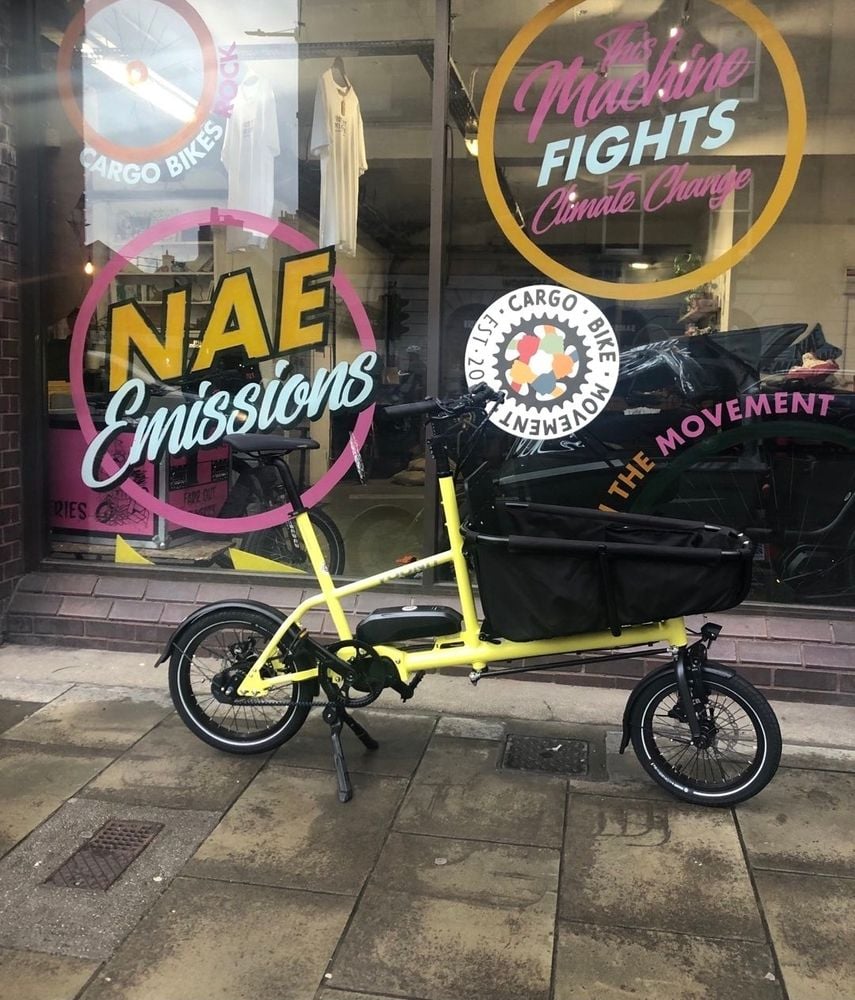 A nimble yellow electric cargo bike with a black front box/basket and small wheels sits in front of the windows of the Cargo Bike Movement hub in Tollcross, Edinburgh. Signs in the window read 'Nae emissions', 'Cargo Bikes Rock', 'Join the Movement' and 'This Machine Fights Climate Change'