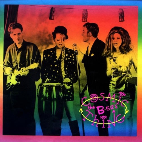 A photograph of the B-52's, tinted with a rainbow of colors, red to yellow to green to blue. They are arranged from left to right, in performance. First is Keith Strickland, a young white man with longish New Wavey hair, playing guitar and smiling. Second is Cindy Wilson, a young white woman wearing a bowler-shaped hat and a very long braid going down her shoulder to her waist. She wears a sparkly coat and is playing a bongo and singing. Third is Fred Schneider, a young white man with short hair, in a suit and tie, holding a microphone in a stand, and facing Cindy, singing to her. Last but not least is Kate Pierson, who has very big red hair that frames her face like a circle. She is wearing a shimmering dress and is singing into a microphone on a stand, and is facing (a presumed) audience.