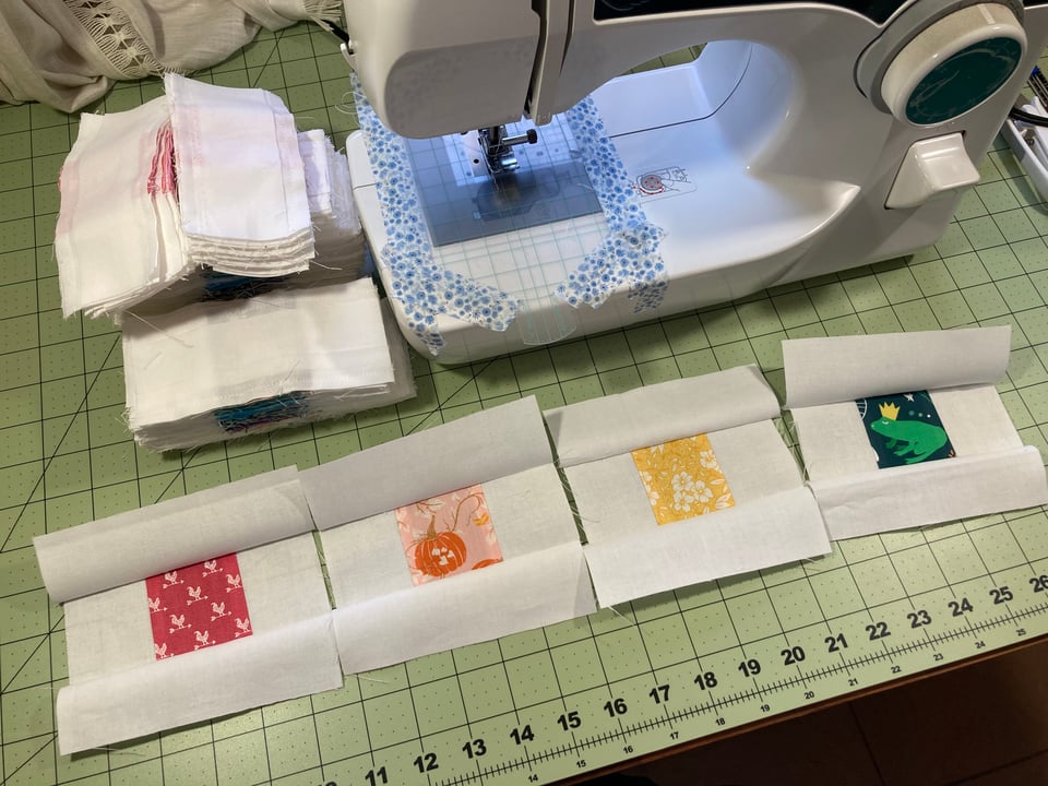 Four pieced-together blocks pulled out in front of a sewing machine to show the colorful centers. They have not been ironed flat yet. The blocks show a center square of fabric, each a different color and pattern, surrounded by white on the top, bottom, and sides, forming a square. From left to right, the patterns show: a red background with white chickens; a pink and orange background with pumpkins and vines; a yellow background with white flowers; and a green background with a tiny frog wearing a yellow crown and puckering up for a kiss.