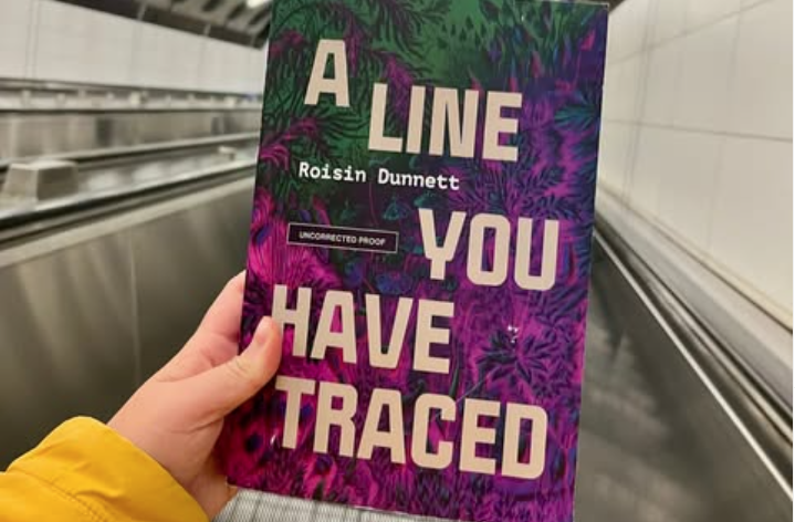 A hand poking out of a yellow sleeve holds the cover of A Line You Have Traced by Roisin Dunnett on an escalator or a moving sidewalk lined with white tiles. The book cover is purple with psychedelic flower/leaf shapes.