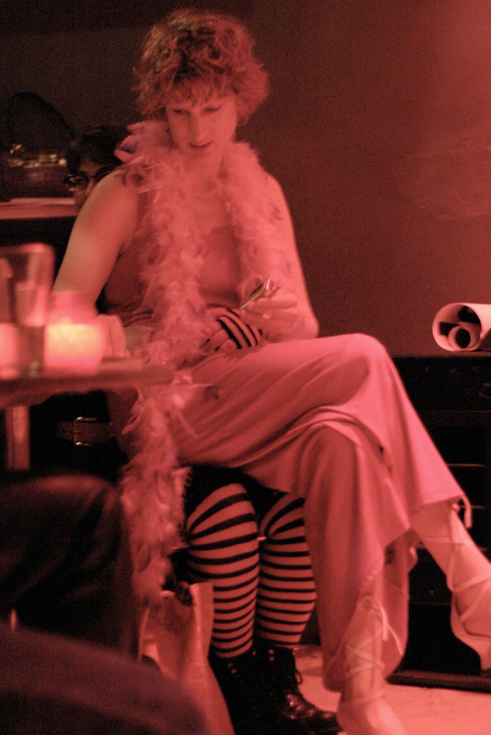 A white trans woman with curly hair wearing a long floofy dress and feather boa, plus high heel shoes with laces going around her calves and ankles, sitting on someone's lap