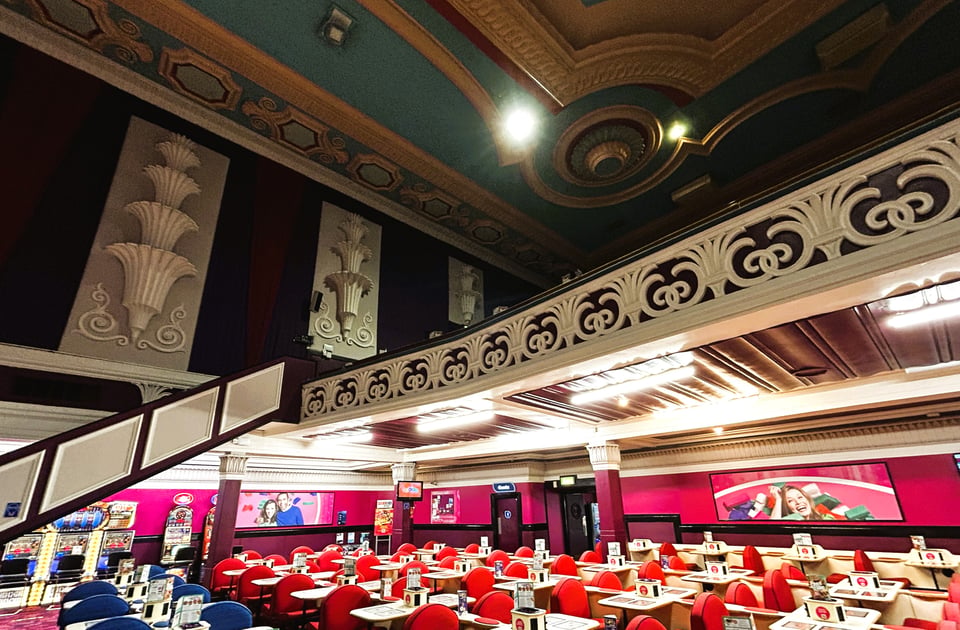 The interior of a former cinema. The balcony is still in place, and has a railing of stylized palm leaves. On the side wall are sculptures of fountains that get smaller as they go up the slope. The ceiling can just be glimpsed in its egg blue and gold glory. In the stalls, bingo tables are in rows.